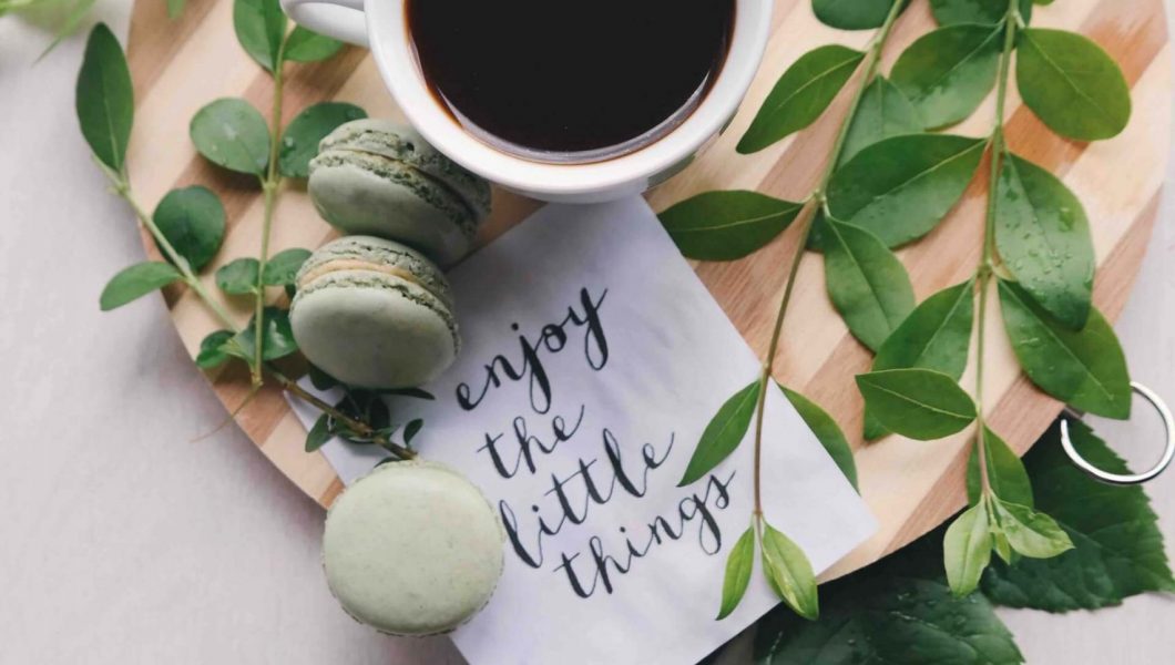 Coffee and macarons on top of a tray with plant branches and a napkin that says "enjoy the little things"