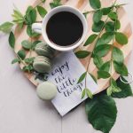Coffee and macarons on top of a tray with plant branches and a napkin that says "enjoy the little things"