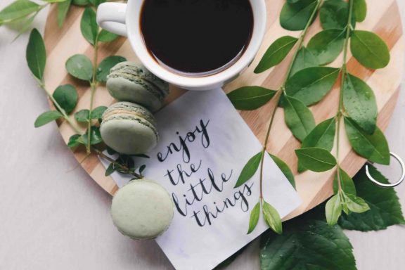 Coffee and macarons on top of a tray with plant branches and a napkin that says "enjoy the little things"