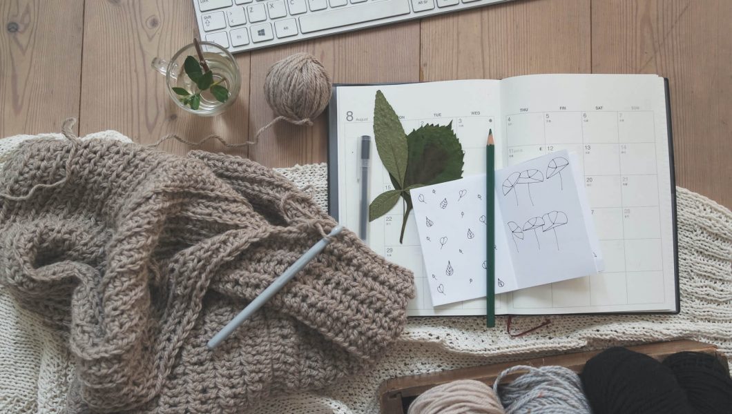 A calendar and sketchpad on top of a desk surrounded by knitting, tea, and a keyboard