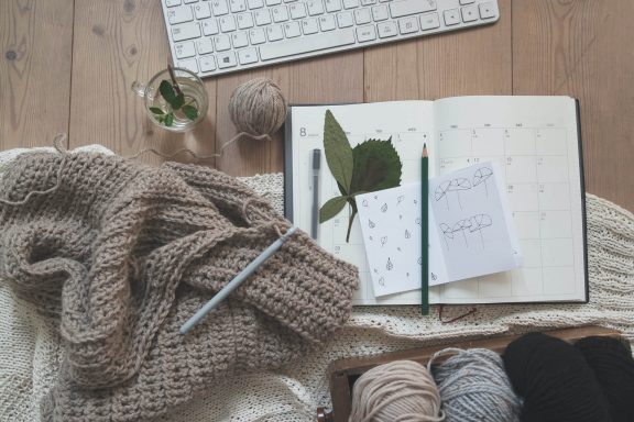 A calendar and sketchpad on top of a desk surrounded by knitting, tea, and a keyboard