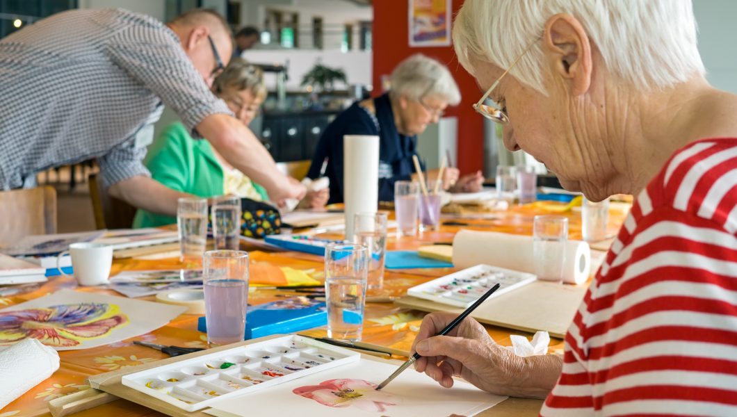 Woman in striped red and white shirt working on watercolor painting at table with other students in spacious studio.