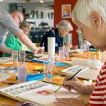 Woman in striped red and white shirt working on watercolor painting at table with other students in spacious studio.