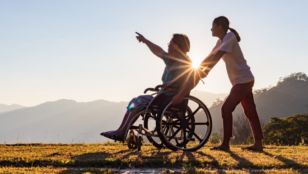 Disabled handicapped young man in wheelchair walking with his care helper in sunset.Silhouette