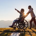 Disabled handicapped young man in wheelchair walking with his care helper in sunset.Silhouette