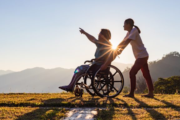 Disabled handicapped young man in wheelchair walking with his care helper in sunset.Silhouette