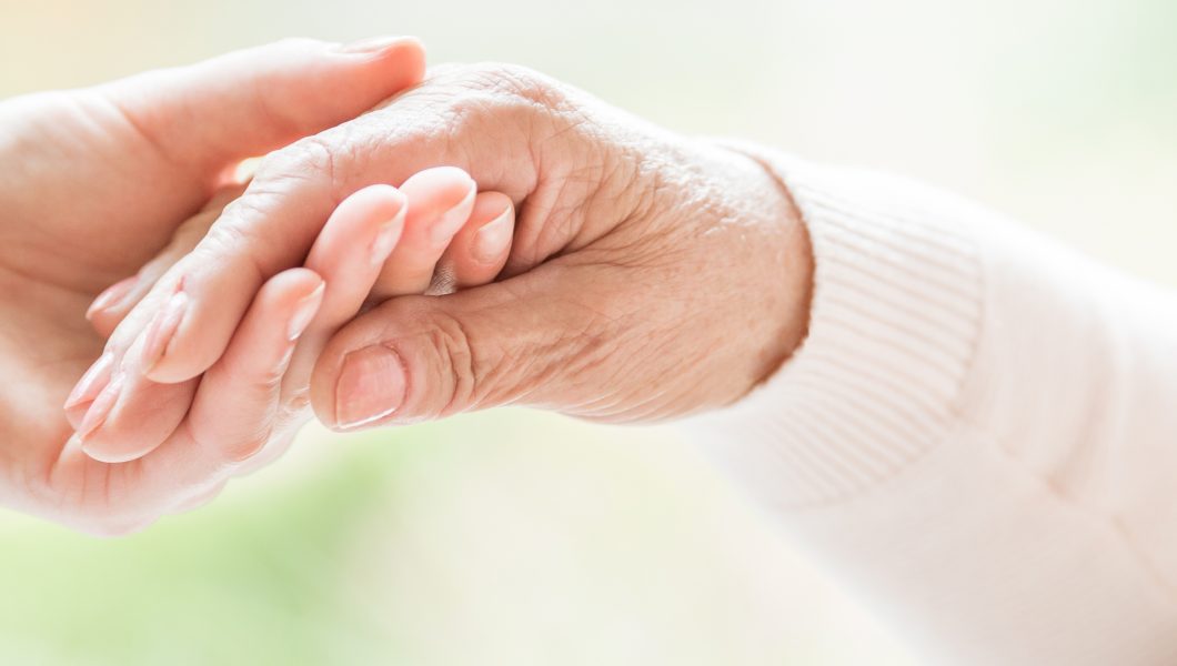 Close-up of tender gesture between two generations. Young woman holding hands with a senior lady. Blurred background. Panorama.