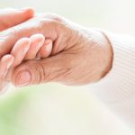 Close-up of tender gesture between two generations. Young woman holding hands with a senior lady. Blurred background. Panorama.