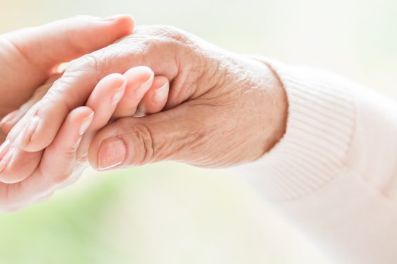 Close-up of tender gesture between two generations. Young woman holding hands with a senior lady. Blurred background. Panorama.